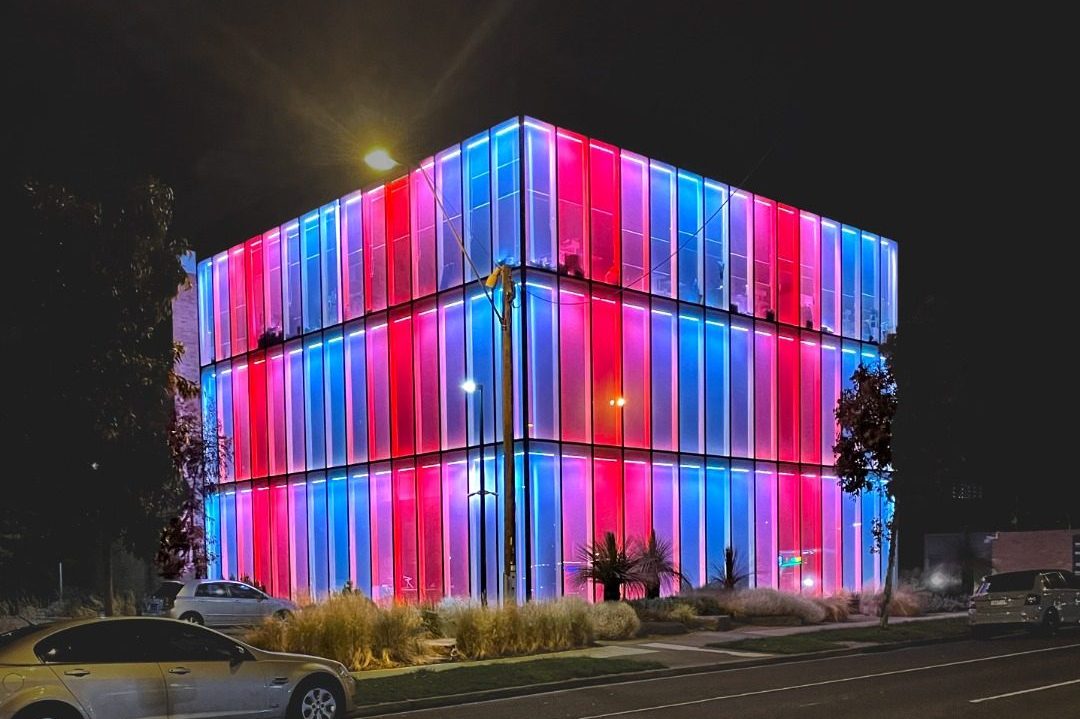 Broadmeadows Town Hall glass cube building is seen from across the street illuminated in red, pink and blue