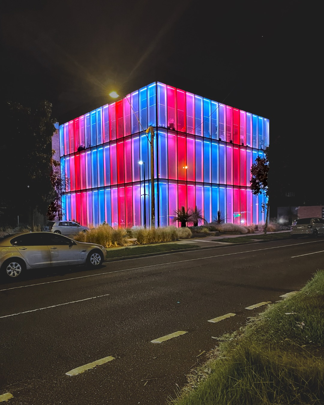 Broadmeadows Town Hall glass cube building is seen from across the street illuminated in red, pink and blue